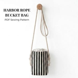 May include: A black and white striped bucket bag with a rope handle, hanging from a wooden peg on a white wall. The bag has a drawstring closure. Text on the image reads: HARBOR ROPE BUCKET BAG PDF Sewing Pattern.
