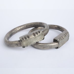 May include: Two silver-toned metal bracelets with a rounded oval shape. Each bracelet has a rectangular clasp with a decorative notched edge. The bracelets are displayed against a white background.