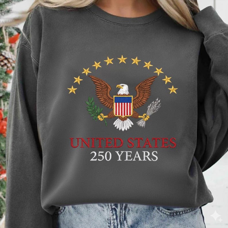 Peut inclure: Sweat-shirt gris fonc&eacute; avec un motif patriotique. Le motif comprend un aigle brun avec un bouclier rouge, blanc et bleu, entour&eacute; d'&eacute;toiles jaunes. Le texte "UNITED STATES 250 YEARS" est imprim&eacute; en rouge et blanc.