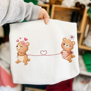 May include: White sweatshirt featuring two cartoon teddy bears connected by a red heart-shaped string. One bear has a pink bow and hearts, the other has a heart-shaped speech bubble. The bears are brown.