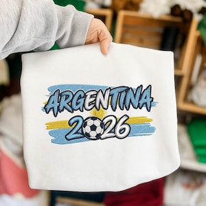 Op de afbeelding: Witte sweatshirt met het woord "ARGENTINA" in blauwe letters boven een voetbal en het nummer "2026". Het ontwerp bevat blauwe en gele penseelstreken, die op een vlag lijken. De sweatshirt wordt door een persoon vastgehouden.