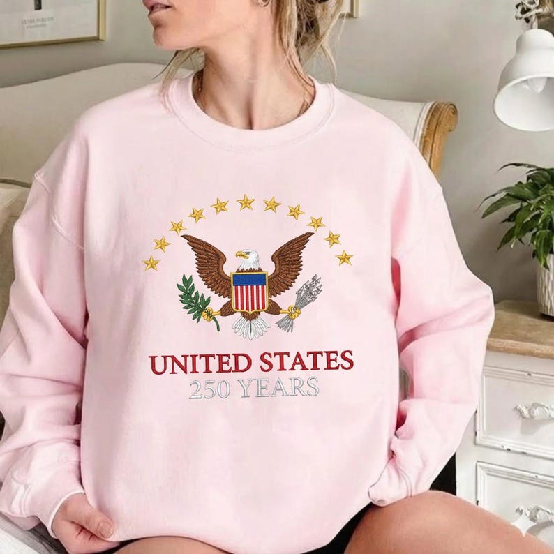 Peut inclure: Un sweat-shirt rose clair avec un motif d'aigle tenant une branche d'olivier et des fl&egrave;ches, entour&eacute; d'&eacute;toiles dor&eacute;es. Le texte "UNITED STATES 250 YEARS" est imprim&eacute; en dessous.