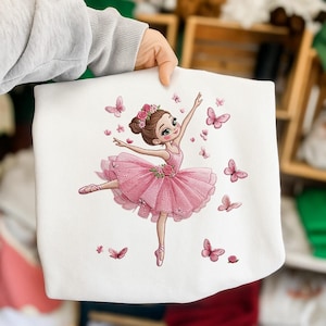 May include: White fabric featuring a ballerina in a pink tutu, arms outstretched, surrounded by pink butterflies. The ballerina has a floral hairpiece. The design is a whimsical illustration, perfect for children's apparel or decor.