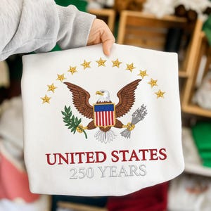 Peut inclure: Sweat-shirt blanc avec un motif brod&eacute;. Il pr&eacute;sente un aigle brun, un bouclier avec des rayures rouges et bleues et des &eacute;toiles dor&eacute;es. Le texte "UNITED STATES 250 YEARS" est brod&eacute; en rouge et gris.