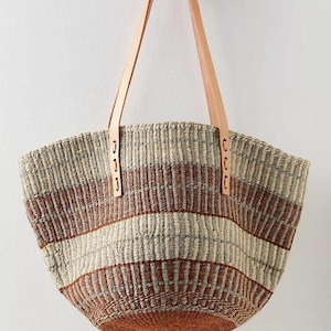 Könnte beinhalten: Eine geflochtene Korb-Tragetasche mit horizontalen Streifen in Beige, Grau und Rostbraun. Die Tasche verfügt über zwei hellbraune Ledergriffe, die mit sichtbaren Nähten befestigt sind, und eine breite, offene Form mit rundem Boden.