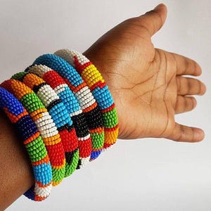May include: A collection of beaded bracelets in various colors, including blue, orange, green, red, yellow, black, and white. The bracelets are stacked on a wrist, showcasing the intricate beadwork and vibrant design. The bracelets appear to be handmade.