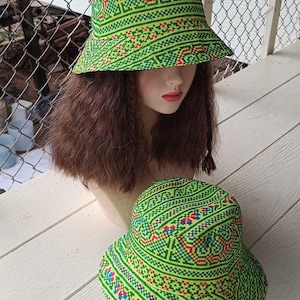 May include: Two green bucket hats with a colorful geometric pattern. The hats have a wide brim and a structured crown. The pattern includes red, blue, and yellow accents on a green background. One hat is on a mannequin head.