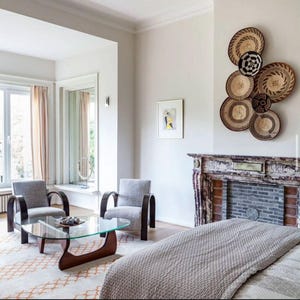 May include: A modern living room with two gray armchairs, a glass-top coffee table, and a decorative wall display of woven baskets. The room features a fireplace and a large window with sheer curtains.