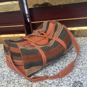 May include: A duffel bag featuring a woven textile body with horizontal stripes in shades of brown, tan, and dark gray. The bag includes tan leather handles, leather trim, and an adjustable leather shoulder strap with antique brass hardware.
