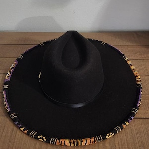 May include: Black fedora-style hat with a wide brim. The brim is decorated with a beaded pattern in shades of purple, orange, and yellow. The hat sits on a wooden surface.