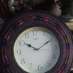 May include: A round wall clock with a white face and black hands. The clock's bezel is decorated with a beaded pattern in shades of pink, purple, and gold. The clock face has black numerals and the brand name "RIKON" is printed at the top.