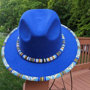 Puede incluir: Un sombrero fedora de color azul real vibrante con una banda de cuentas alrededor de la copa y un borde de cuentas a juego a lo largo del ala. El intrincado trabajo de cuentas incorpora patrones en tonos azul claro, blanco, amarillo y negro.