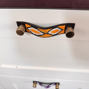 Può includere: Primo piano di un mobile bianco con una maniglia decorativa. La maniglia presenta una base in pelle marrone scuro con un design di perline arancioni, bianche e nere. Le manopole sono dorate.
