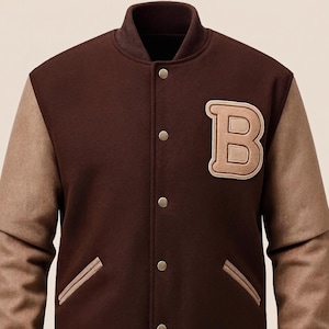 Op de afbeelding: Een donkerbruin varsity jack met beige mouwen en een geribbelde kraag, boorden en zoom. De voorzijde is voorzien van een grote beige letter B-patch op de linkerborst, twee zijzakken met beige biezen en een rij zilverkleurige metalen drukknopen.
