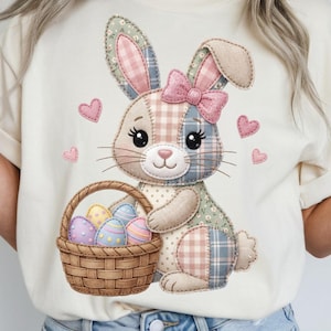Può includere: T-shirt color crema con un coniglietto patchwork che tiene un cestino di uova di Pasqua. Il coniglietto ha un fiocco rosa ed è circondato da cuori rosa. Il cestino contiene uova dai colori pastello, perfetto per Pasqua.