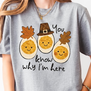 May include: A light gray t-shirt featuring a graphic of three smiling egg characters with fall-themed decorations. One egg wears a pilgrim hat. The text "You know why I'm here" is printed below the eggs.