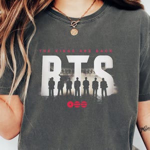 May include: Dark gray t-shirt with the text "THE KINGS ARE BACK" above the white letters "BTS". Below the letters is a silhouette of seven figures and three red circles. The shirt is worn with blue jeans.