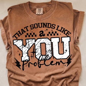 May include: A copper-colored short-sleeve t-shirt with a distressed graphic print. The text reads THAT SOUNDS LIKE a YOU problem in black and white varsity-style lettering, accented with small stars, checkerboard patterns, and lightning bolts.