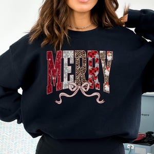 May include: Black sweatshirt with the word "MERRY" in a festive design. The letters feature patterns like red and white squares, snowflakes, leopard print, and Santa Claus faces. A bow is below the word.