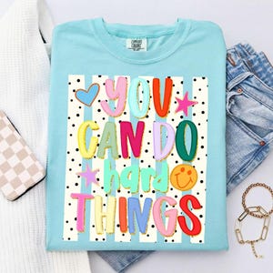 May include: A light blue Comfort Colors t-shirt with the phrase "YOU CAN DO hard THINGS" in colorful, bubble letters. The design includes a blue heart, stars, and a smiling face. The background has vertical stripes and black polka dots.
