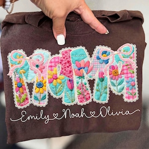 May include: A brown sweatshirt with the word "MOM" appliqued in colorful floral patterns. The letters are outlined with white lace and topped with pink bows. Below the word "MOM" are the names "Emily Noah Olivia" in a cursive font.