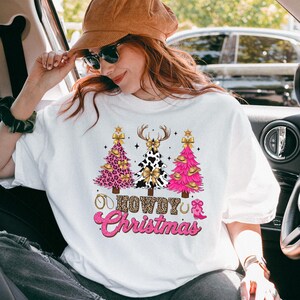 May include: White crew neck sweatshirt with a festive "Howdy Christmas" design. The graphic features three Christmas trees with cow print, pink leopard print, and pink tinsel. The text is in a pink, glittery font.
