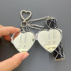 Puede incluir: Llavero con forma de corazón plateado con cierre en forma de corazón. El relicario se abre para revelar dos superficies espejadas. Un dije cilíndrico negro y plateado está sujeto al llavero mediante una cadena.