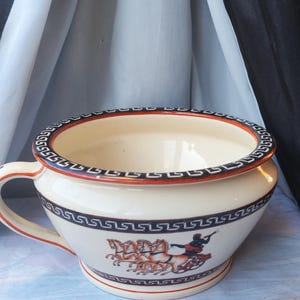 Puede incluir: Orinal de cerámica color crema con asa. El orinal presenta un diseño de borde rojo y negro y una escena de carro griego. El interior es blanco y el orinal se asienta sobre una pequeña base.
