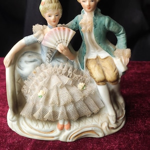 Könnte beinhalten: Porzellanfigur, die ein Paar in Kleidung des 18. Jahrhunderts darstellt. Die Frau sitzt und hält einen Fächer, während der Mann neben ihr steht. Die Figur hat Pastellfarben, filigrane Spitzendetails und einen dekorativen Sockel.