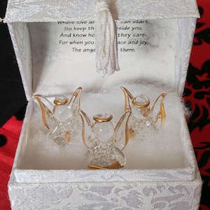Op de afbeelding: Drie heldere glazen engelfiguurtjes met goudkleurige halo's en vleugelaccenten, geplaatst in een met witte stof beklede doos. Het deksel heeft een wit kwastje en de tekst: Where love and life can start, So keep this angel beside you, And know how much they care, For when you need peace and joy, The angel will be there.