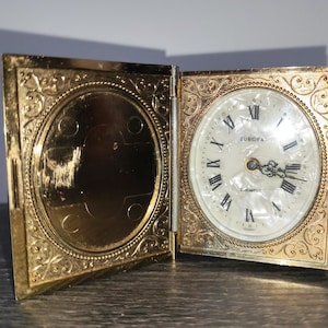 May include: A gold-colored, folding travel clock with an ornate design. The clock face is white with black Roman numerals and the word "EUROPA". The clock is open, revealing the time.