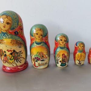 Könnte beinhalten: Ein Satz von fünf russischen Matrjoschka-Puppen, jede bemalt mit einer anderen Szene und einem Porträt einer Frau. Die Puppen sind hauptsächlich rot, gelb und blau, mit aufwendigen Details und einem glänzenden Finish. Die größte Puppe ist etwa 15 cm groß.