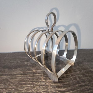 May include: A silver-colored, vintage toast rack with a handle. The rack has a series of curved dividers, designed to hold slices of toast upright. The design is simple and functional, with a classic aesthetic. The rack is made of metal and sits on a dark surface.