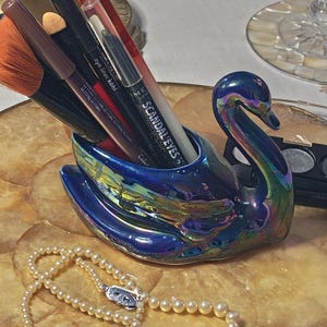 Könnte beinhalten: Ein schwanenförmiger Halter mit irisierendem blau-violettem Finish, der mehrere Make-up-Pinsel und Eyeliner-Stifte enthält, darunter einer mit der Aufschrift Rimmel London Scandaleyes. Eine Kette aus weißen Perlen liegt vor der Schwanenfigur.