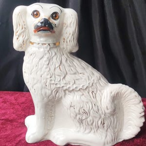 Op de afbeelding: Een witte keramische hondenfiguur met bruine ogen, een zwarte neus en gouden accenten op de oren en halsband. De hond zit, met een pluizige, getextureerde vacht. De figuur staat op een rode ondergrond.