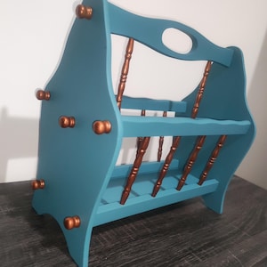 May include: A teal wooden magazine rack featuring a central handle cutout and copper-colored spindle accents. The structure includes two tiers for storage, with small copper-colored knobs decorating the exterior side panels of the frame.