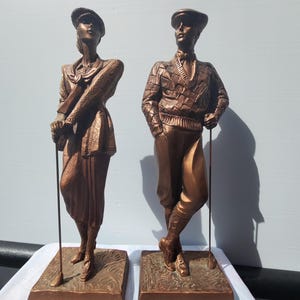 May include: A pair of bronze-toned statues depicting two golfers standing on rectangular bases. One figure wears a collared jacket and skirt, while the other wears a patterned sweater and knickerbockers. Both figures hold golf clubs.