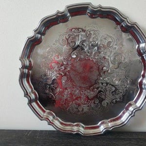 Peut inclure: Un plateau de service rond de couleur argentée avec un bord festonné et une surface réfléchissante. Le centre et la bordure présentent des volutes en relief complexes et des gravures florales créant un motif texturé sur le métal.