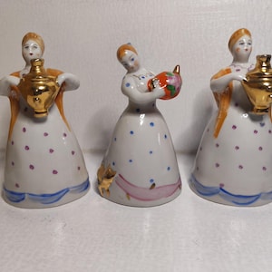 Puede incluir: Tres figuras de porcelana de mujeres con trajes tradicionales. Cada figura es blanca con una falda estampada y un samovar o tetera dorada. Una figura sostiene una tetera colorida. Las figuras tienen detalles en naranja y dorado.