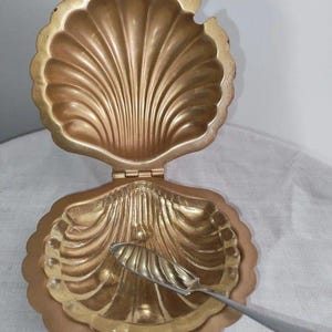 May include: A gold-toned metal clam shell dish with a hinged lid that opens to reveal a scalloped interior. A small, silver-toned metal serving spoon with a twisted handle rests inside the bottom half of the shell dish.
