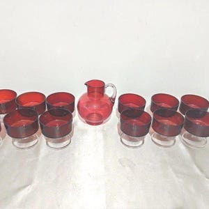 Puede incluir: Un juego de doce cuencos de postre de cristal rojo rubí y una jarra de cristal a juego. Cada cuenco presenta un recipiente rojo intenso apoyado sobre una base de cristal transparente. La jarra central tiene cuerpo redondeado, asa y pico.
