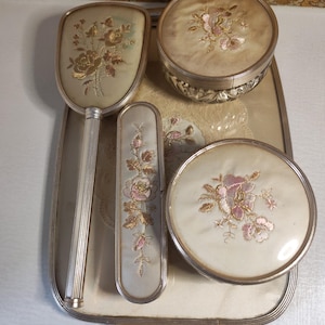 Könnte beinhalten: Ein Vintage-Schminkset mit Handspiegel, zwei runden Behältern und einem langen Pinsel, alles auf einem silberfarbenen Tablett. Das Set zeigt gestickte Blumenmuster in Rosa, Gold und Grün auf cremefarbenem Hintergrund. Wahrscheinlich aus der Mitte des 20. Jahrhunderts.