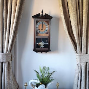 May include: A dark wood wall clock with a light-coloured clock face and Roman numerals hangs on a white wall. Below, a white swan-shaped planter holds greenery, flanked by two brass candle holders. Beige patterned curtains frame the scene.
