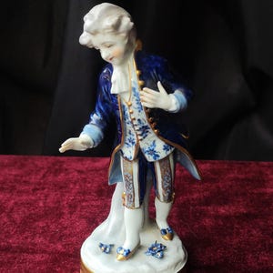 Può includere: Statuina in porcellana di un uomo in abiti del XVIII secolo. La figura indossa una giacca blu con finiture dorate, un gilet bianco con motivi floreali blu e calzoni bianchi. La base è bianca con dettagli dorati e floreali blu.
