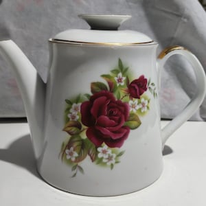 Può includere: Teiera in ceramica bianca con coperchio e manico. La teiera è decorata con una grande rosa bordeaux e fiori bianchi più piccoli con foglie verdi. Il bordo e il manico hanno dettagli dorati.
