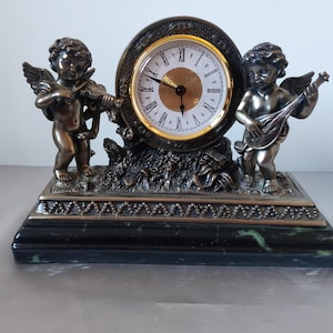 Op de afbeelding: Een decoratieve klok met bronzen cherubs, een die viool speelt en de andere mandoline, die een wijzerplaat met Romeinse cijfers flankeren. De klok heeft een zwart-groene marmeren basis en sierlijke details.
