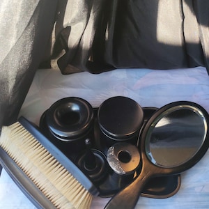 Op de afbeelding: Een vintage zwarte toilettafelset met een handspiegel, borstel en diverse containers. De set bevat een dienblad, een borstel met lichtgeleurde borstelharen en verschillende kleine potjes. De spiegel heeft een donkerhouten handvat.