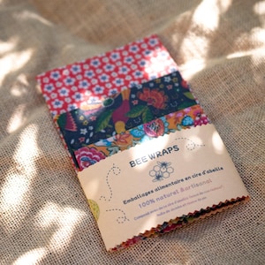 May include: A stack of rectangular beeswax food wraps featuring various floral and botanical patterns in red, navy blue, and mustard yellow. The wraps are secured with a tan paper band labelled BEE WRAPS with bee and honeycomb graphics.