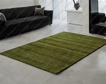 May include: A rectangular, olive green rug with a shaggy texture. The rug is placed on a light-colored tiled floor, with a black sofa and other furniture visible in the background. The rug's dimensions are not visible.
