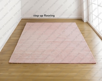 Op de afbeelding: Een rechthoekig, lichtroze tapijt op een houten vloer. Het tapijt is gecentreerd in een kamer met witte muren en een raam. De tekst "step up flooring" is zichtbaar.